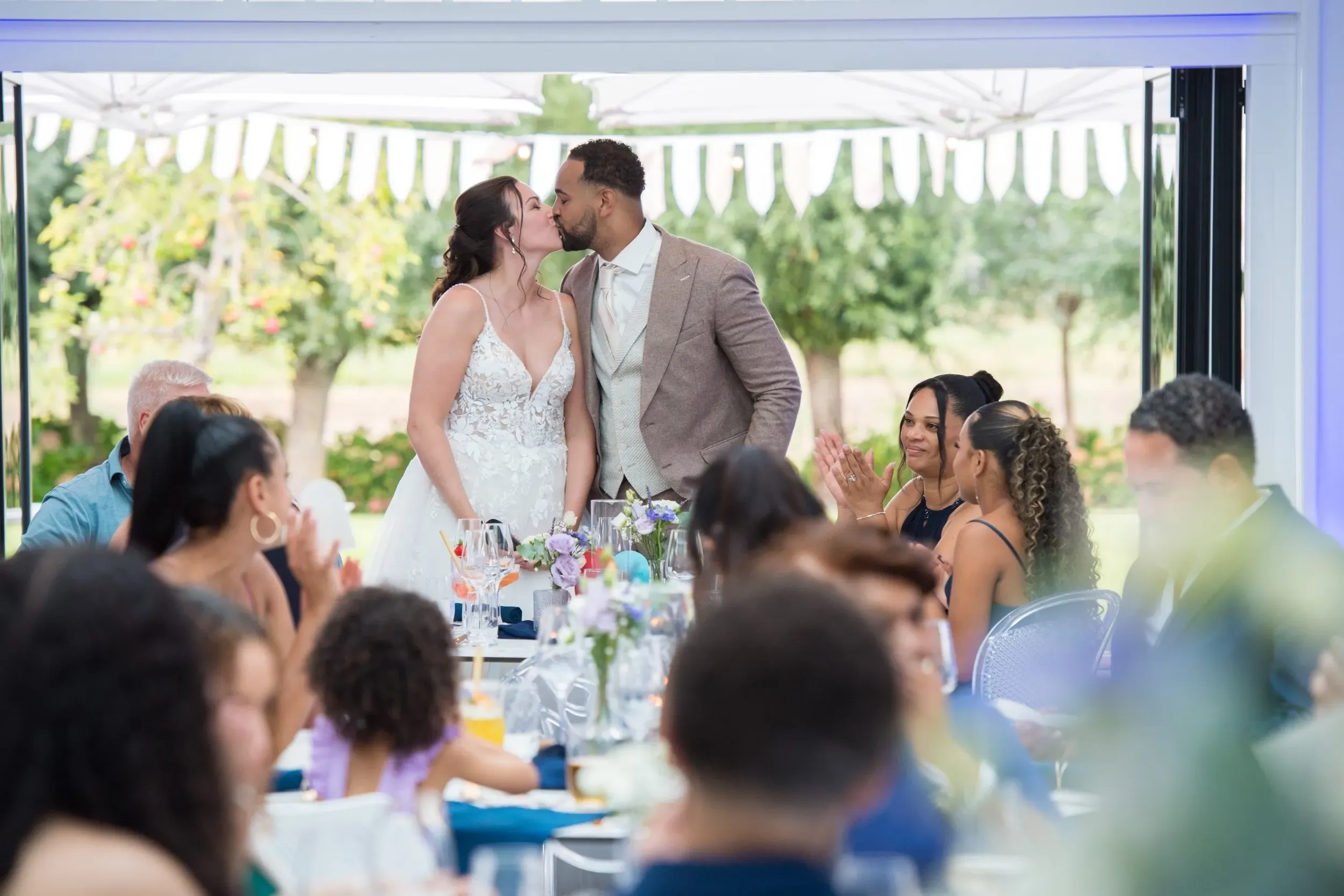Een pasgetrouwd stel in bruidskleding deelt een kus, omringd door hun dierbaren in de Orangerie, waar de deuren openstaan naar de groene tuin.