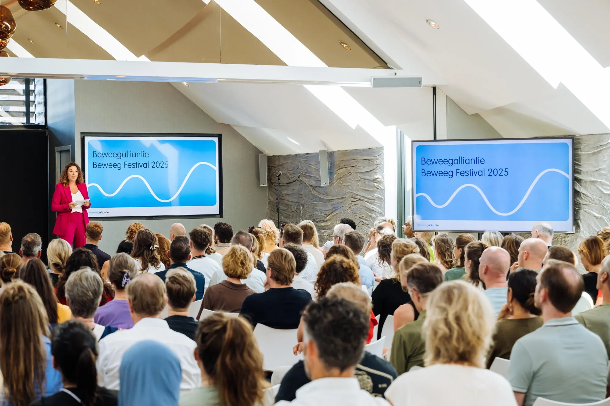 Een zakelijke bijeenkomst waar gasten in theateropstelling aandachtig naar een spreker luisteren die haar verhaal toelicht via een presentatie op twee grote led-schermen.
