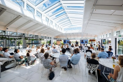Een zakelijke bijeenkomst in de open, met daglicht gevulde orangerie waar gasten aan de ronde tafels zitten en aandachtig luisteren naar een spreker die voorin tussen twee grote LED-schermen staat.