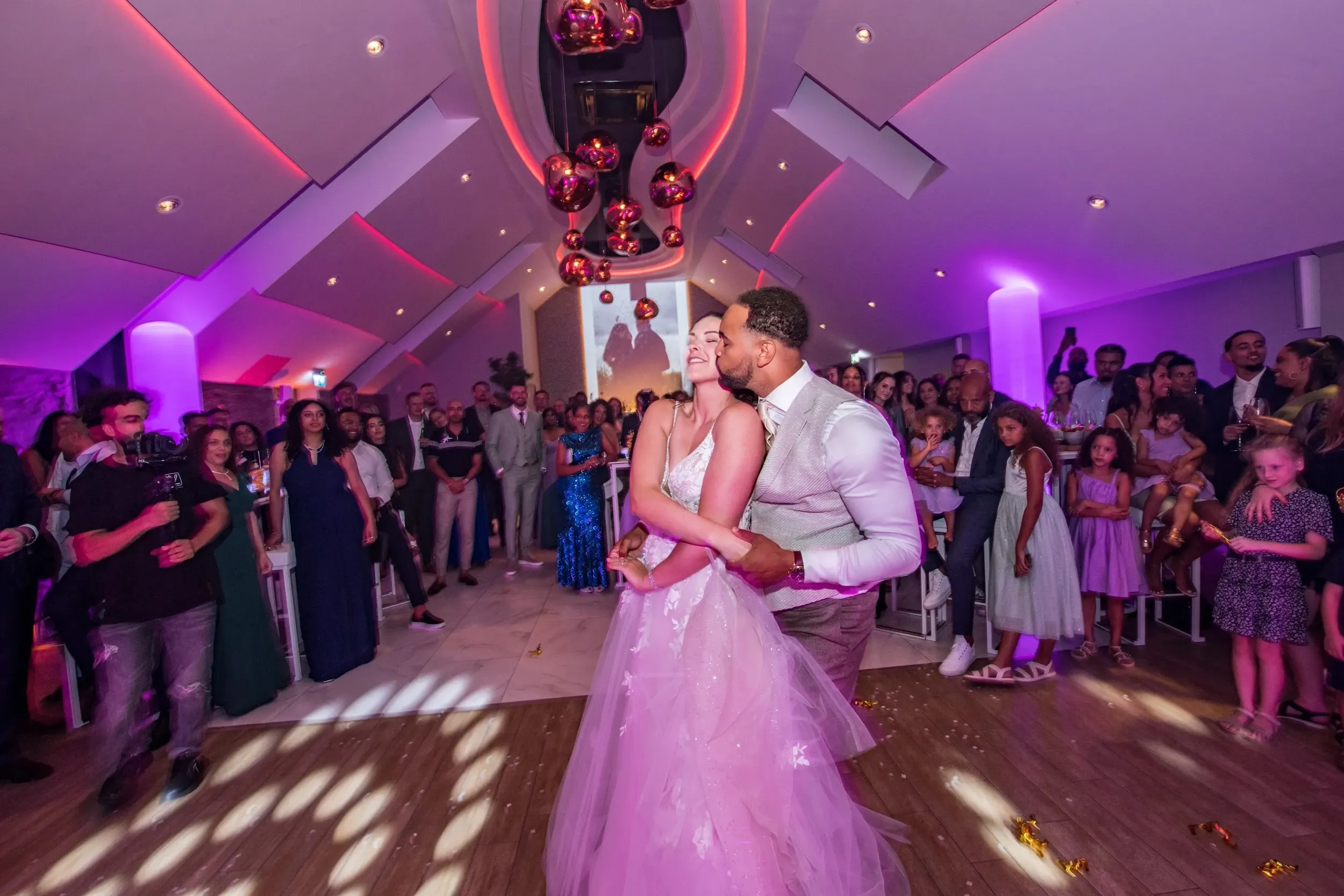 Een bruidspaar danst intiem op de dansvloer van de feestzaal met romantische sfeerverlichting en gasten die naast de vloer hun moment aanschouwen.