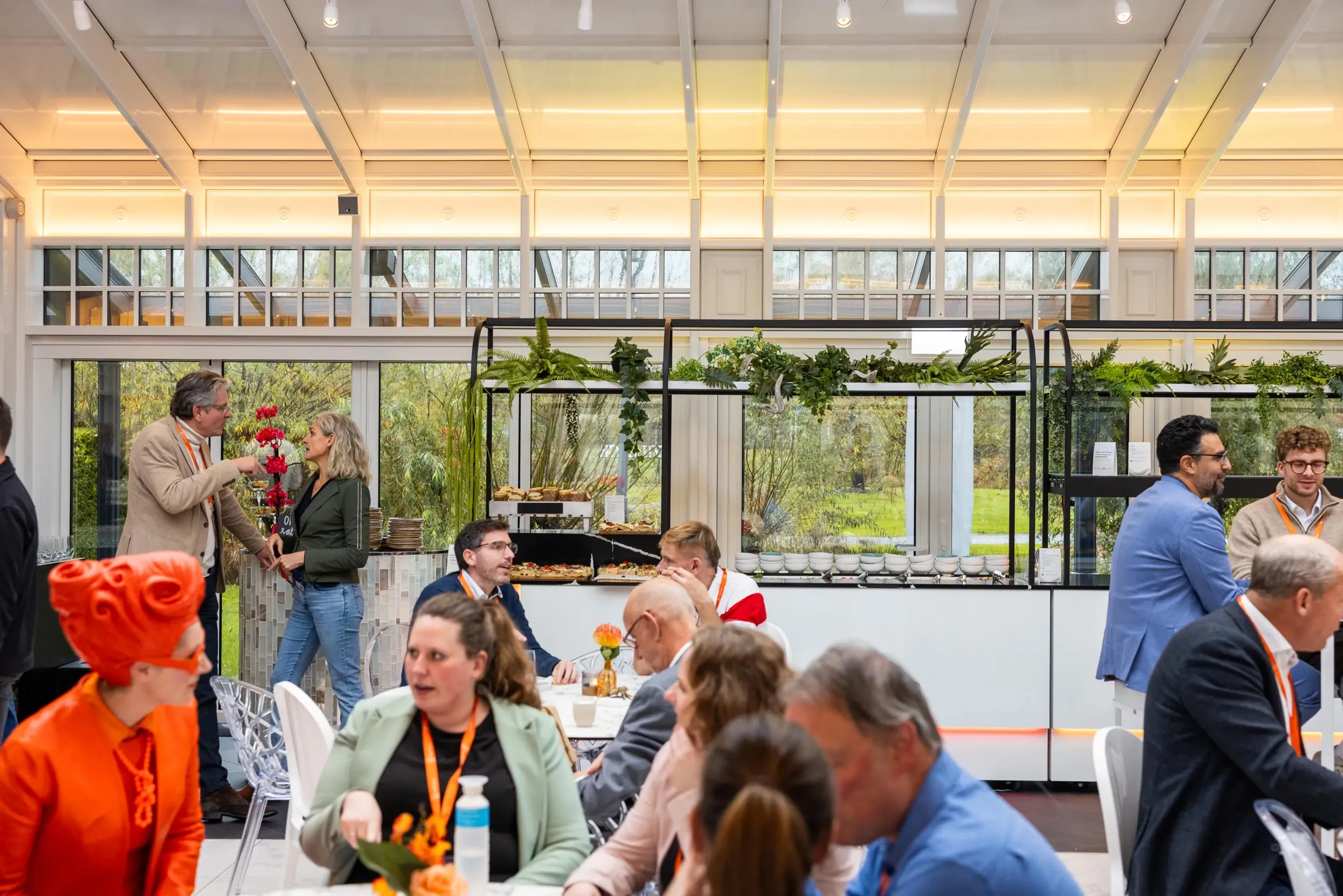 Gasten zijn in gesprek aan ronde tafels in een ruimte met greote ramen waar ook een opstelling voor een buffet klaarstaat.