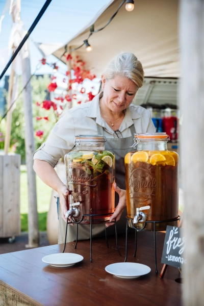 Een medewerker van Green Village in een schort plaatst glimlachend een grote glazen drank dispenser met verse ijsthee op een tafel van het buffet.